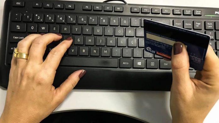 Mulher com uma mão sobre teclado e outra segurando cartão de débito e crédito