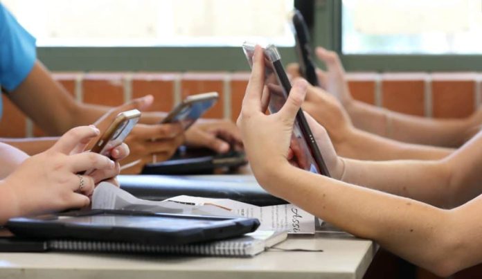 Mesa com alunos do ensino médio, todos segurando vários dispositivos digitais, como telefones e tablets, durante uma aula.