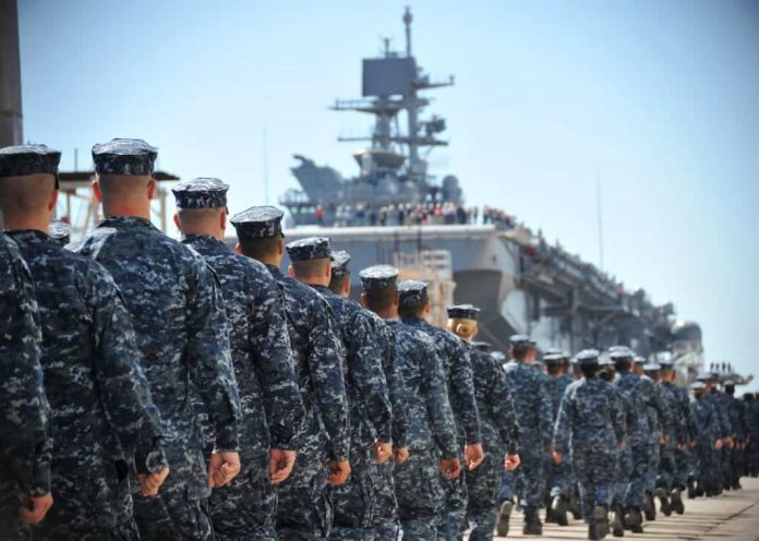 Imagem mostra marinheiros e fuzileiros navais dos EUA designados para o navio de assalto anfíbio Pre-Commissioning Unit America (LHA 6) marcham em direção ao navio no píer da Huntington Ingalls Industries em Pascagoula.