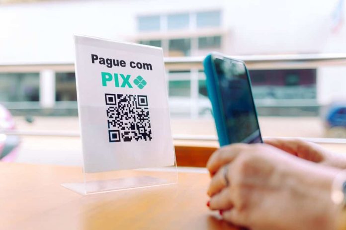 QR Code com os dizeres "pague com Pix" acima; ao lado, uma pessoa posiciona um smartphone para ler o código