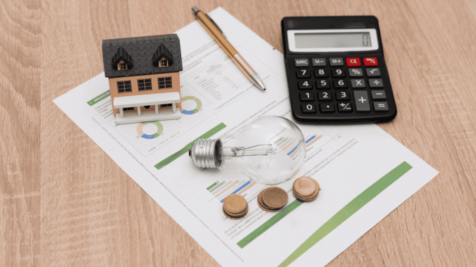imagem mostra um boleto/ conta de energia elétrica em cima de uma mesa madeira com calculadora, lâmpada, moedas e a miniatura de uma casa