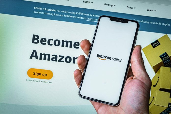 Ao fundo, site do Amazon Seller emum monitor; à frente, mão de uma pessoa segurando um smartphone com o logo do Amazon Seller